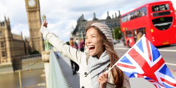 London - happy tourist holding British UK flag by Big Ben and red double decker bus. Excited girl sightseeing travel on Westminster Bridge, London, England, United Kingdom. Multiracial Asian Caucasian